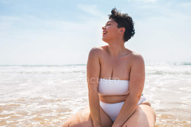 Sorridente giovane plus size femminile in costume da bagno seduto sulla spiaggia sabbiosa guardando lontano vicino all'oceano schiumoso sotto il cielo nuvoloso blu alla luce del giorno — Foto stock