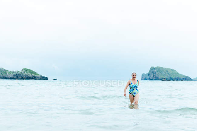 Positive active fit aged female in swimsuit walking out of sea water while enjoying summer day on beach — Stock Photo