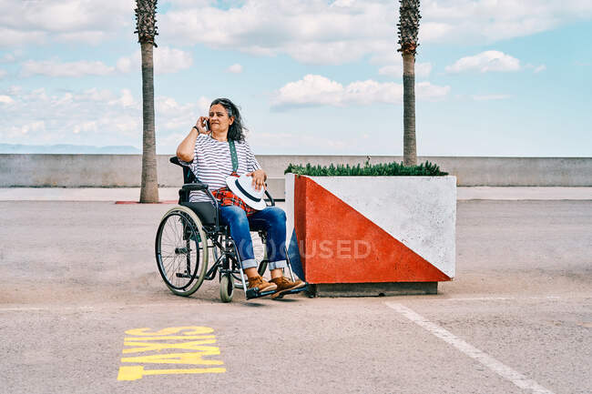 Зріла жінка з обмеженими можливостями сидить у інвалідному візку і розмовляє на мобільному телефоні, викликаючи таксі на парковці біля набережної — стокове фото