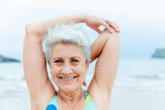 Положительный седой пенсионерка в купальнике растягивая плечи во время фитнес-тренировки на пляже возле размахивая морем в летний день — стоковое фото