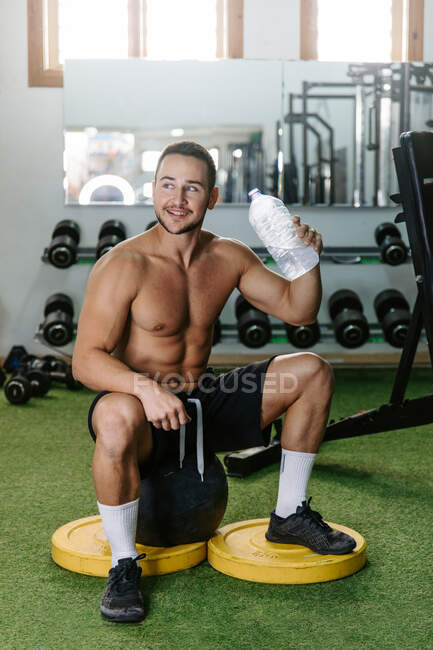 Deportista sentado en el gimnasio moderno y disfrutando de agua dulce durante el entrenamiento después de hacer ejercicio - foto de stock