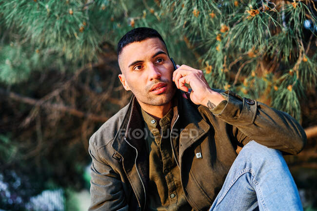 Hispanic guy in casual clothes looking away and answering phone call while standing near barrier and coniferous tree in park — Stock Photo