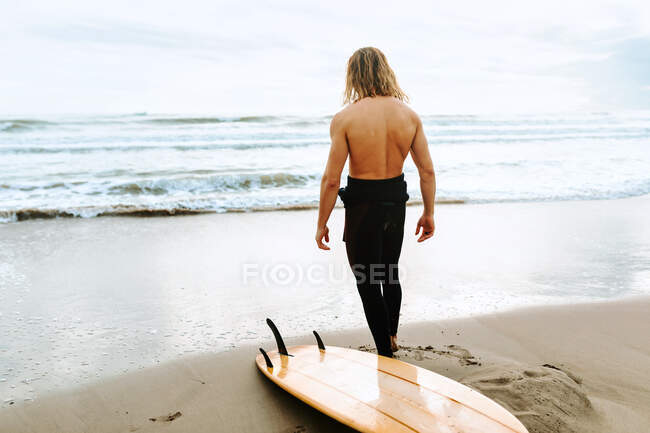 Vista posterior de surfista irreconocible hombre con el pelo largo vestido con traje de neopreno de pie con tabla de surf en la playa - foto de stock