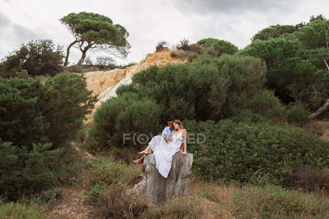 Мультирасовая пара молодоженов сидит на пне дерева в лесу и обнимается в день свадьбы — стоковое фото