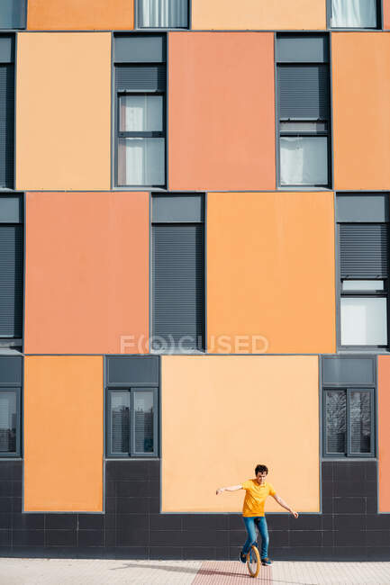 Full body of young guy in casual wear riding on unicycle on modern urban street with colorful building — Stock Photo