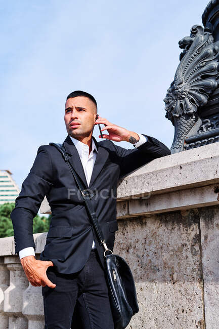 Впевнений молодий чоловік в елегантному формальному костюмі говорить на мобільному телефоні і посміхається, стоячи біля скульптури на міській площі — стокове фото