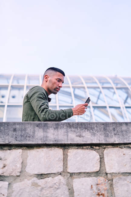 Low angle of adult Hispanic man in shirt leaning on stone wall and browsing cellphone outside contemporary glass building — Stock Photo