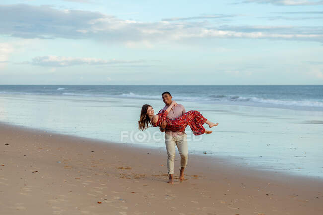 Happy black man carrying delighted woman and enjoying summer on sandy seashore on background of sea at sunset — Stock Photo