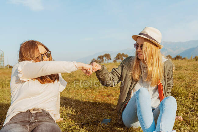 Close girlfriends in hat and sunglasses sitting on lawn in mountains and bumping fists in sunlight — Stock Photo
