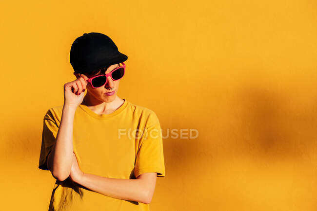 Confident female in black cap looking away and adjusting vivid pink sunglasses against yellow background — Stock Photo