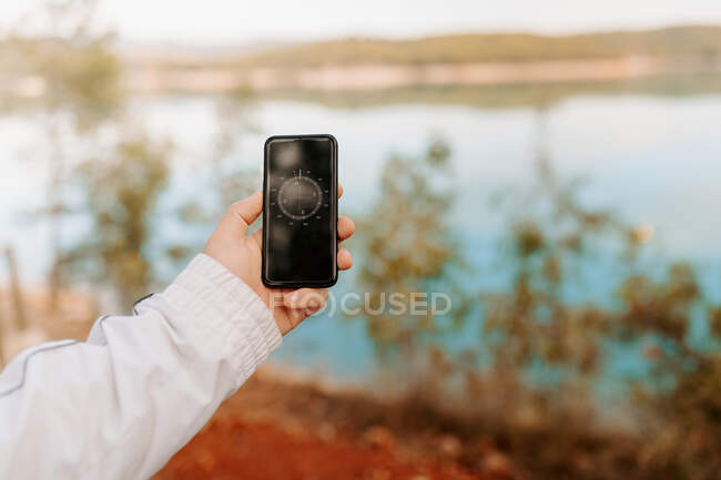 Recortado irreconocible persona sosteniendo el teléfono móvil mientras mira la aplicación brújula en medio de un bosque con lago - foto de stock
