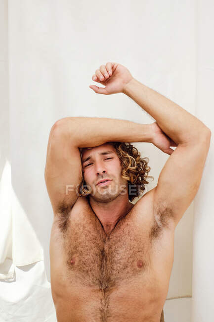 Handsome male with naked torso sitting on background of white curtain and looking at camera — Stock Photo