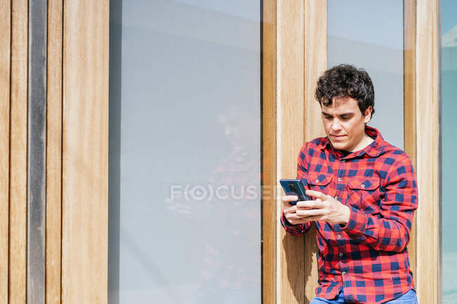 Молодий чоловік у повсякденному носінні використовує смартфон, стоячи проти сучасної міської будівлі — стокове фото
