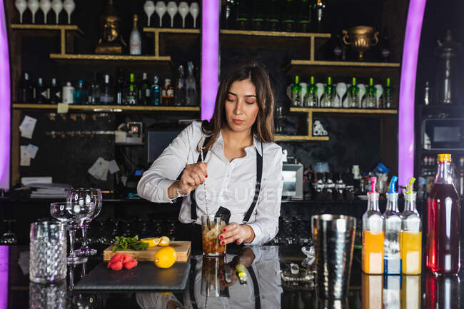 Female barkeeper in stylish outfit adding ice cubes into glass and stirring with long spoon while preparing cocktail standing at counter in modern bar — Stock Photo