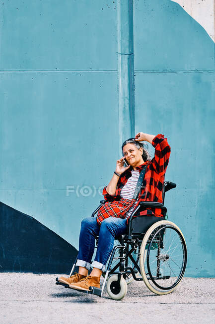 Selbstständige behinderte ältere Frau im Rollstuhl telefoniert in der Nähe einer bemalten Wand auf der Straße — Stockfoto