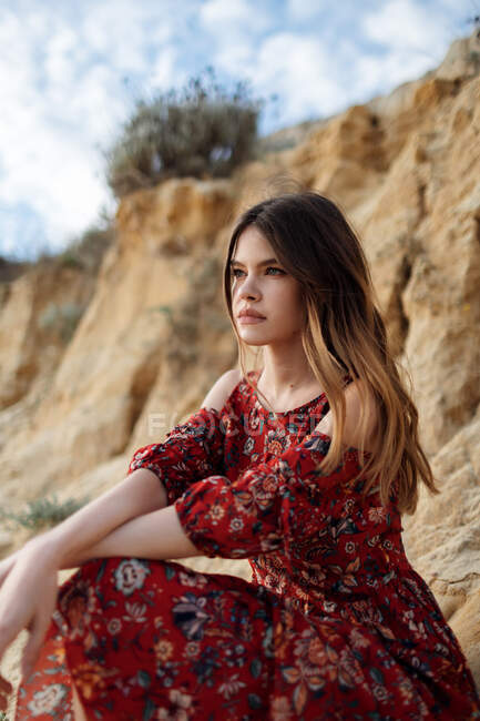Спокойная женщина в длинном летнем платье сидит на песчаном холме и смотрит в сторону — стоковое фото