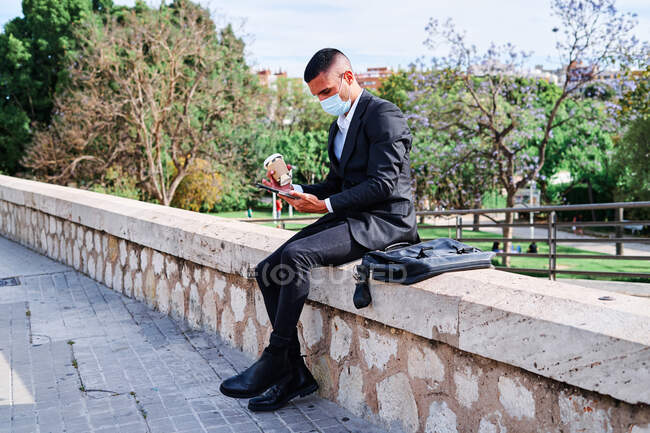 Вид сбоку на концентрированного молодого бизнесмена в элегантном костюме и защитной маске, читающего новости на планшете во время разрыва с чашкой кофе на городской улице во время пандемии коронавируса — стоковое фото