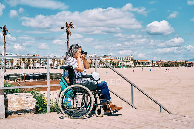 Vista lateral de una mujer discapacitada irreconocible de pelo gris en silla de ruedas tomando fotos con cámara fotográfica mientras pasa el día de verano en la playa - foto de stock