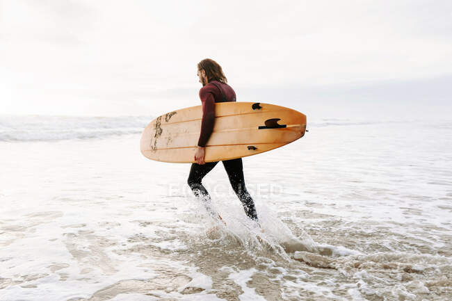 Vista lateral do surfista vestido de fato de mergulho correndo com prancha de surf em direção à água para pegar uma onda na praia durante o nascer do sol — Fotografia de Stock