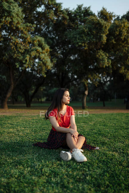 Спокойная женщина, сидящая на траве на лугу в парке и наслаждающаяся закатом летом — стоковое фото