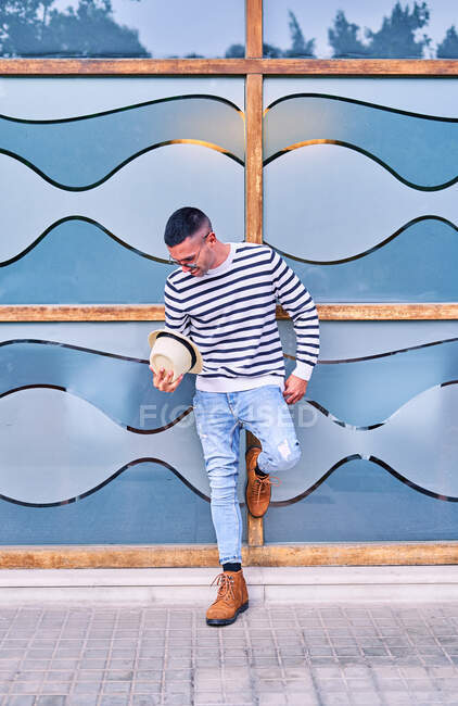 Full body adult Hispanic male in trendy outfit leaning on glass wall with abstract ornament and looking away — Stock Photo