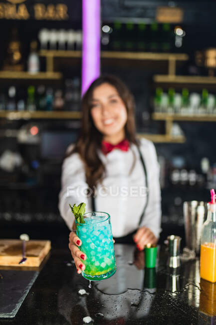 Barman femme méconnaissable floue dans une tenue élégante servant cocktail bleu avec feuille de menthe tout en se tenant au comptoir dans le bar moderne — Photo de stock
