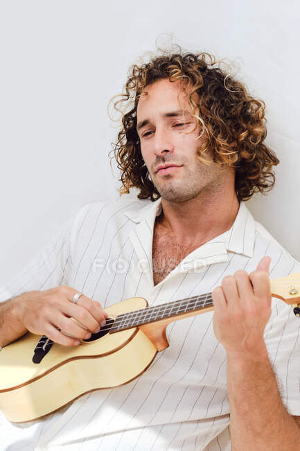 Спокойный мужчина с вьющимися волосами, опирающийся на белую стену и играющий на укулеле, отводя взгляд — стоковое фото
