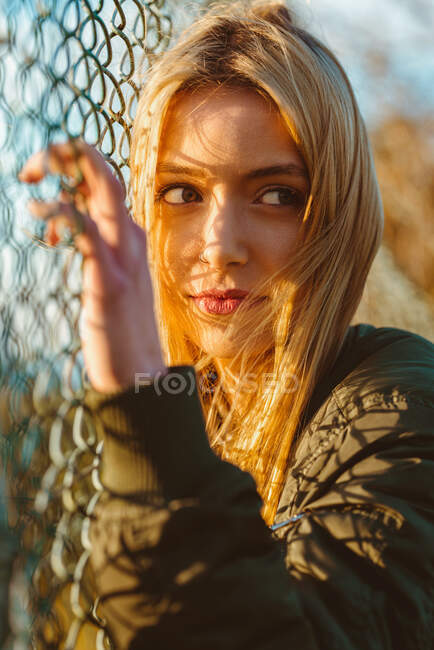 Hermosa hembra rubia con chaqueta mirando hacia otro lado parada en la cerca de eslabones de cadena bajo la luz del sol dorada - foto de stock