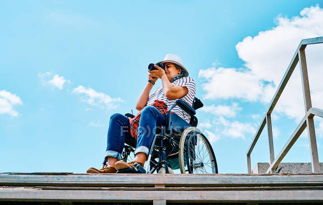 From below gray haired disabled woman in wheelchair taking pictures on photo camera near stairway against blue sky in city — Stock Photo