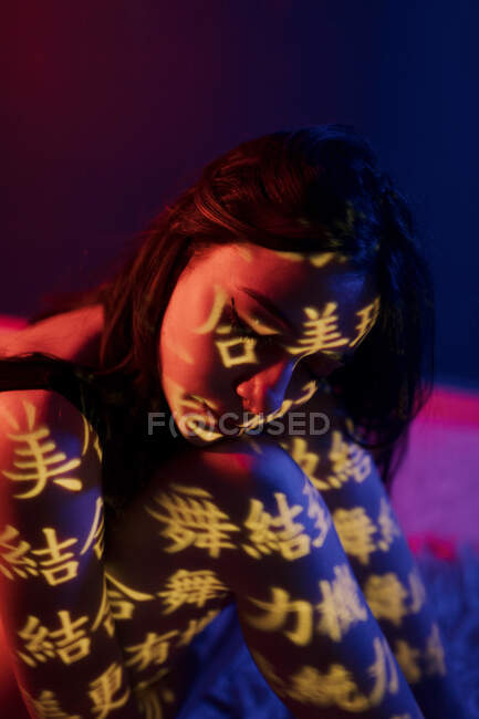 Fashionable young female model with light projection in shape of oriental hieroglyphs sitting with eyes closed leaning on knees in dark studio with red illumination — Stock Photo