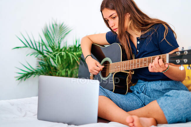 Estudante focada assistindo vídeo tutorial no laptop enquanto aprendia a tocar guitarra acústica durante o tempo livre em casa — Fotografia de Stock