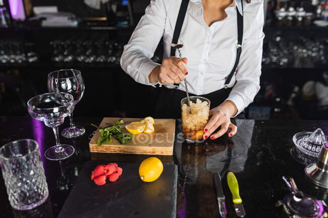 Cropped unrecognizable female barkeeper in stylish outfit adding ice cubes into glass and stirring with long spoon while preparing cocktail standing at counter in modern bar — Stock Photo