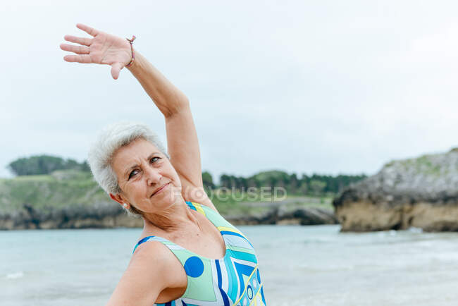 Positive senior female in stylish swimsuit doing side bend exercise while practicing healthy lifestyle and working out on beach against waving ocean — Stock Photo