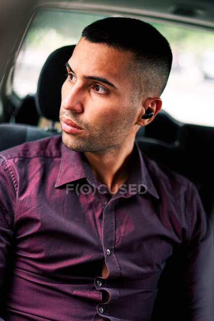 Hombre hispano barbudo joven confiado en camisa elegante usando auriculares inalámbricos verdaderos mientras que está sentado en el asiento del pasajero en auto y mirando hacia otro lado - foto de stock