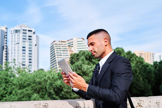 Side view of focused young Hispanic male executive manager in classy suit with bag reading information on tablet while standing near railing on urban terrace — Stock Photo