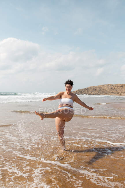 Pleine longueur de la jeune femelle en maillot de bain éclaboussant l'eau de mer tout en se tenant sur la côte sablonneuse par temps ensoleillé sous le ciel nuageux bleu — Photo de stock