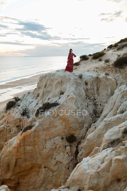 Vue à distance de la femelle en robe debout sur une colline rugueuse sur fond de mer et de coucher de soleil — Photo de stock