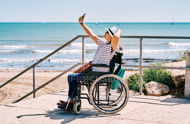 Vue latérale complète du corps du voyageur féminin handicapé positif assis en fauteuil roulant et prenant selfie sur smartphone tout en passant des vacances d'été sur la plage près de l'océan — Photo de stock