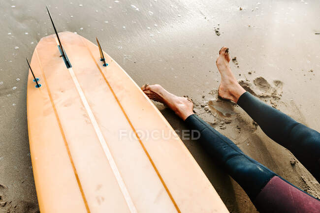 From above cropped anonymous surfer man dressed in wetsuit sitting with surfboard on the beach during sunrise — Stock Photo