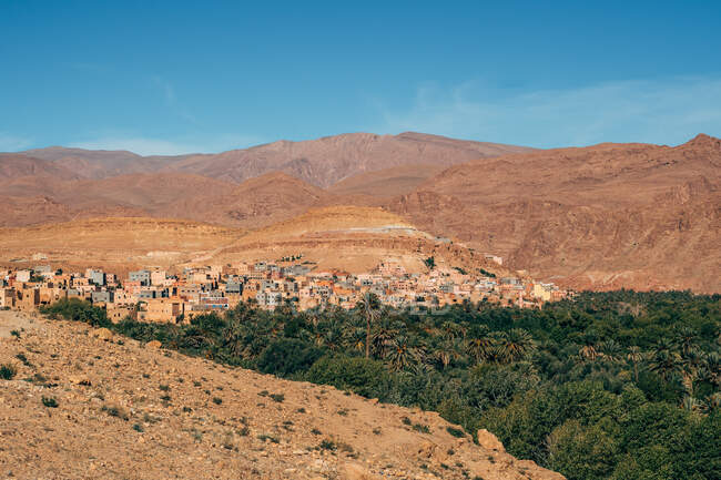 Dall'alto di vecchi edifici in pietra sul pendio di grandi montagne colorate tra piante verdi con cielo azzurro chiaro sullo sfondo in Marocco — Foto stock
