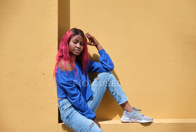 Vista lateral de la confiada adolescente afroamericana con el pelo teñido usando camisa azul brillante y jeans rasgados con zapatillas de deporte sentadas cerca de la pared amarilla y mirando a la cámara - foto de stock