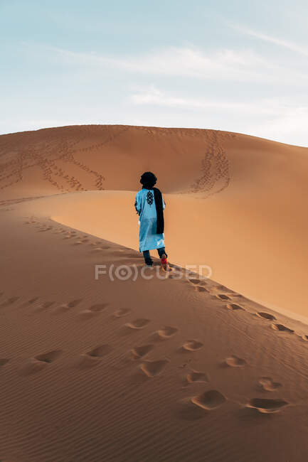 Visão traseira da senhora em vestido azul claro e pano preto na cabeça em pé na duna de areia vazia com céu azul no fundo em Marrocos — Fotografia de Stock