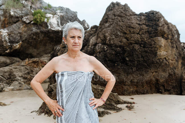 Senior viajera envuelta en toalla de pie en la playa de arena con formaciones rocosas mientras descansa después de nadar en el mar en el día de verano - foto de stock