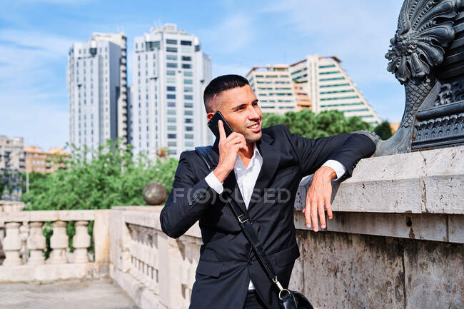 Confident young man in elegant formal suit talking on mobile phone and smiling while standing near sculpture on urban square — Stock Photo