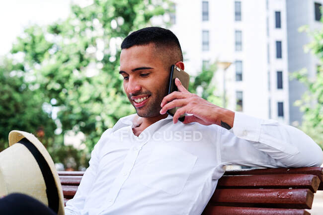 Bajo ángulo de sonriente auto seguro joven barbudo caballero hispano en traje elegante y sombrero hablando en el teléfono móvil mientras descansa en el banco en la ciudad - foto de stock
