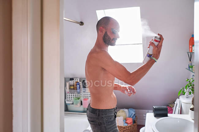 Vue latérale du mâle barbu torse nu avec drag queen maquillage appliquer pulvérisation sur le visage tout en se tenant dans la salle de bain — Photo de stock