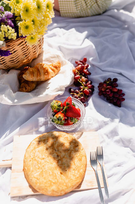 From above of crop faceless woman sitting on blanket with croissant and berries placed near Italian focaccia with wicker basket of blooming flowers — Stock Photo