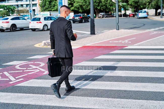 Full body of young well dressed male entrepreneur in classy suit and protective mask carrying briefcase and disposable cup of takeaway drink walking on crosswalk in summer day in city — Stock Photo