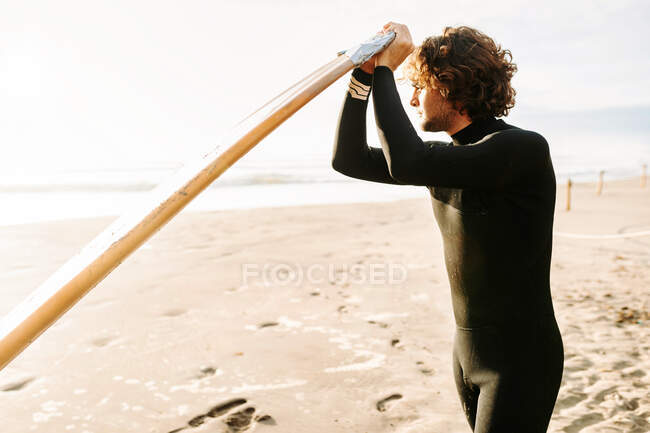 Hombre surfista vestido con traje de neopreno de pie mirando hacia otro lado con la tabla de surf en la playa durante el amanecer en el fondo - foto de stock
