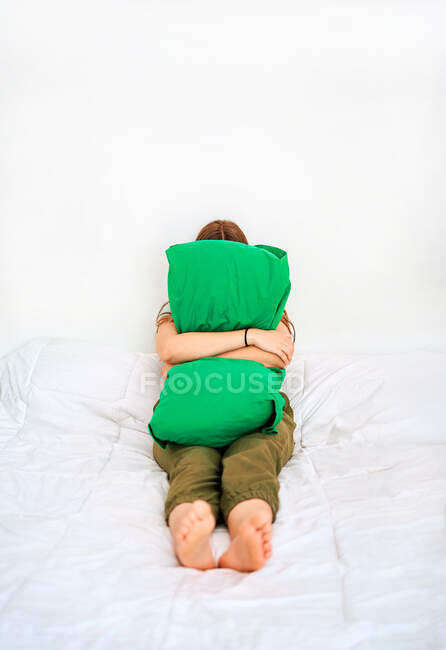 Полная длина неузнаваемой несчастной женщины, обнимающей зеленую подушку, сидя в одиночестве дома на кровати — стоковое фото